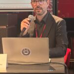 Relatore sul palco durante la presentazione sulla direttiva NIS2, con slide proiettata e pubblico attento.